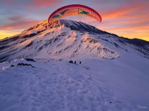 A colorful paraglider soars above a snow-capped mountain during a vibrant sunset, with silhouettes of two people below on the slope.