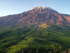 Majestic Mount Kilimanjaro towers over verdant hills, its snow-capped summit illuminated by the sunlight.