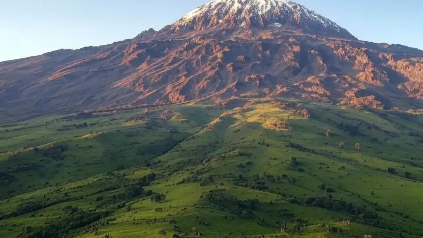 Majestic Mount Kilimanjaro towers over verdant hills, its snow-capped summit illuminated by the sunlight.