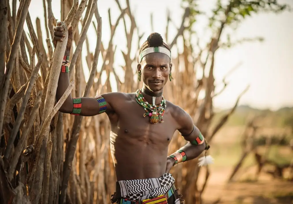 A man from the Hadza tribe with tribal tattoos stands confidently in front of a large tree in a natural setting.