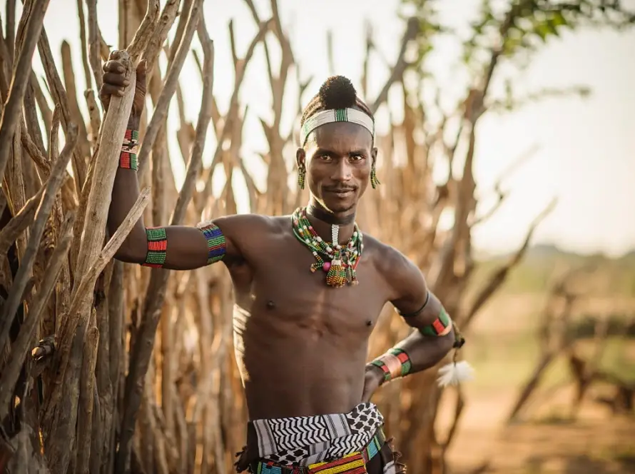 A man from the Hadza tribe with tribal tattoos stands confidently in front of a large tree in a natural setting.