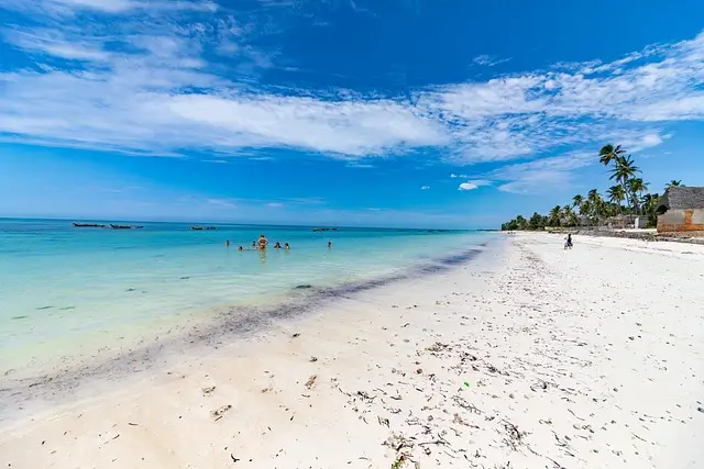 White sandy beach with scattered seaweed, clear blue water, a few people swimming, palm trees, and boats in the distance under a partly cloudy sky.