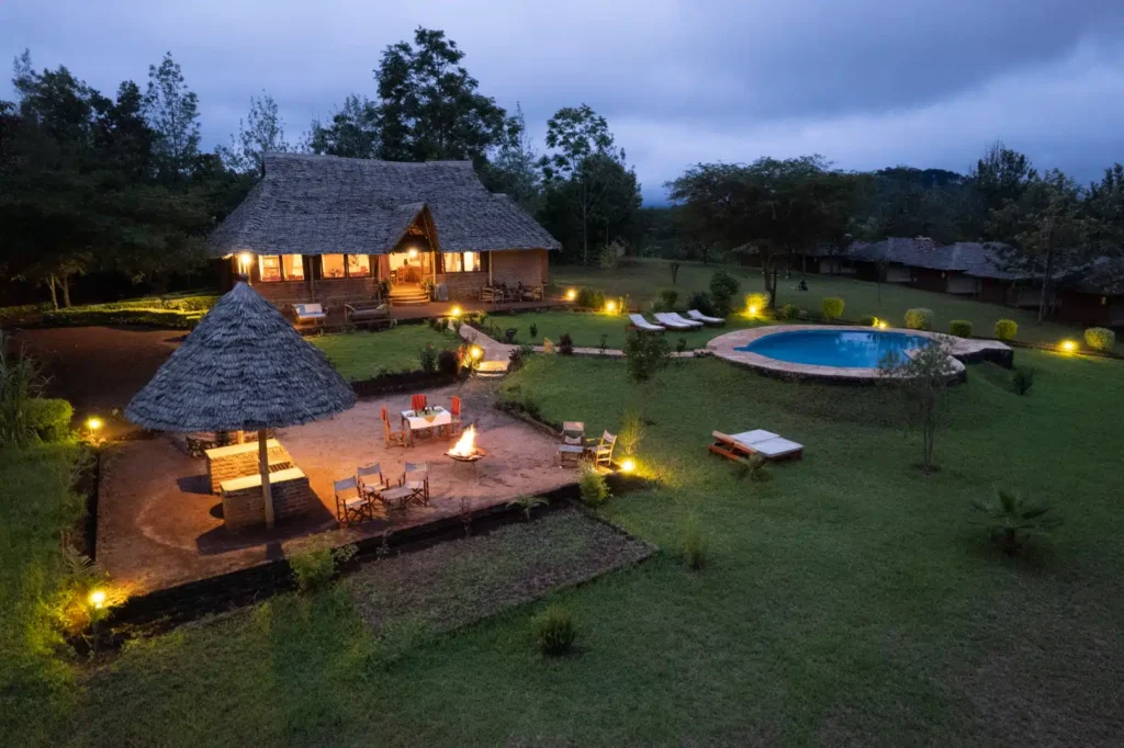 A safari lodge lit up at dusk with a thatched main building, firepit dining area and swimming pool