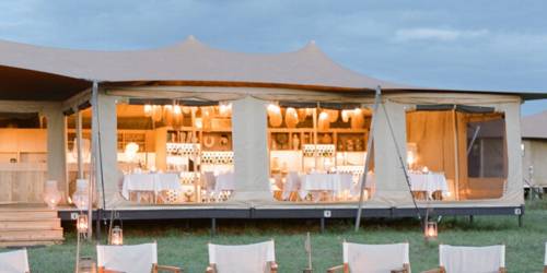 Luxury camping tent with lit interior and campfire, chairs set outside on grass at dusk
