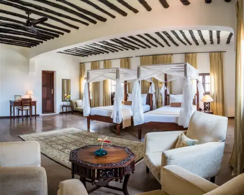 Luxurious hotel room featuring two four-poster beds, elegant decor, plush seating, and rich wooden beams in a bright, airy space