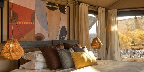 Stylish tent interior featuring a cozy bed with decorative pillows, woven lamps, artwork, and natural light from large windows