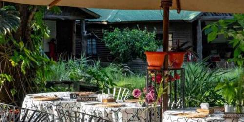 Outdoor cafe setting with patterned tables, black chairs, and a large umbrella amidst greenery at Mount Meru Game Lodge