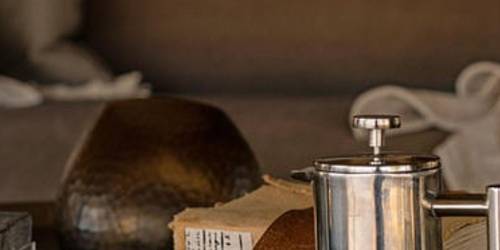 A stainless steel French press beside a white coffee cup on a round table with two stacked books and a decorative sphere in the background