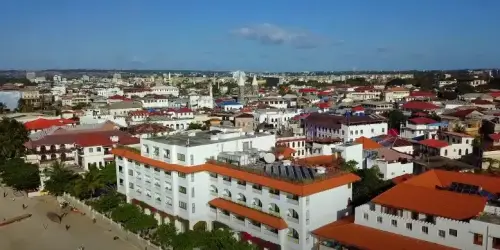 Aerial view of a vibrant coastal city with varied architecture, featuring white buildings with orange roofs and lush greenery