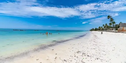 White sandy beach with scattered seaweed, clear blue water, a few people swimming, palm trees, and boats in the distance under a partly cloudy sky.
